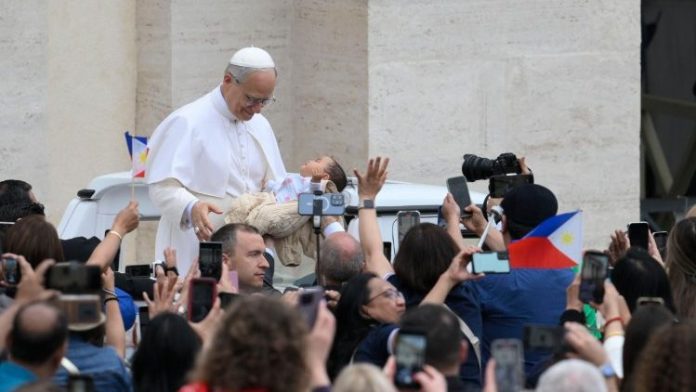 Paus Leo XIV membopong anak kecil dalam perjalanan berkeliling Lapangan Santo Petrus Vatikan, Rabu 21 Mei 2025, sesaat sebelum menggelar audiensi umum pertama kalinya. (Vatican Media)